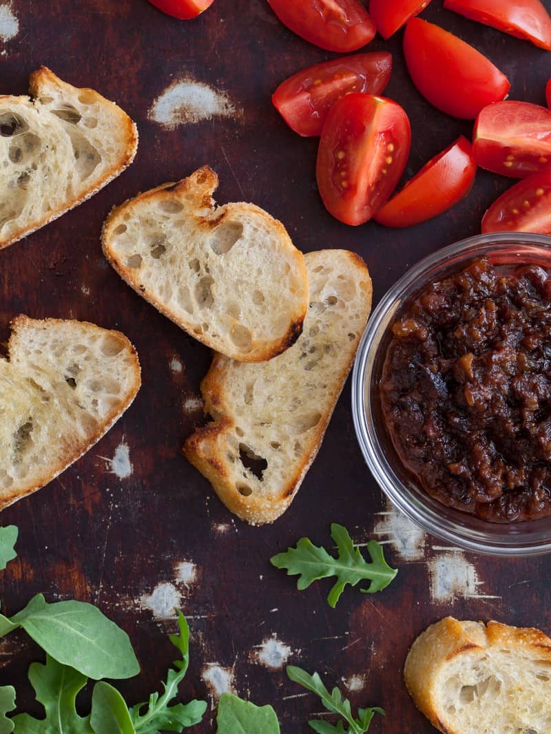 Boozy Bacon Jam recipe A close up of crostini with tomatoe wedges and a bowl of boozy bacon jam.