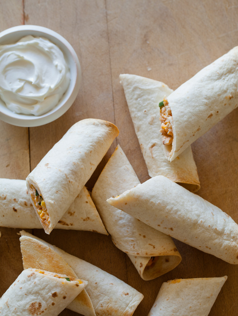 Quick and easy baked chicken flautas with a ramekin of sour cream on a wooden surface.