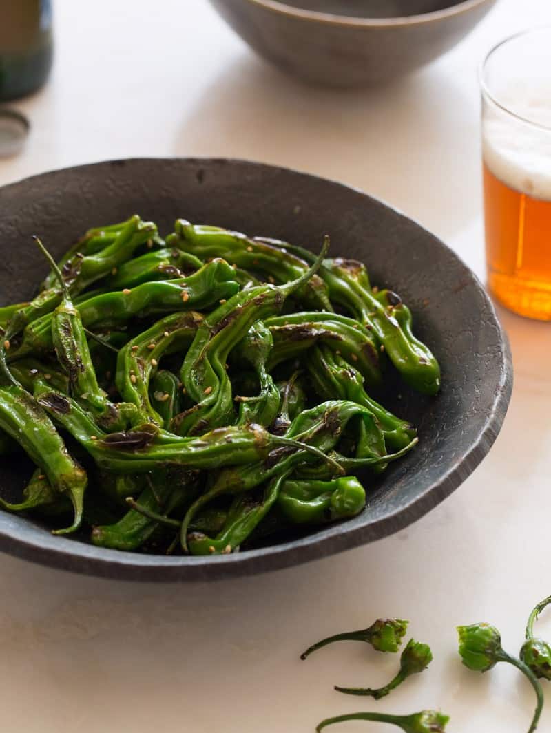 Blistered shishito peppers in a dark bowl next to a beer.