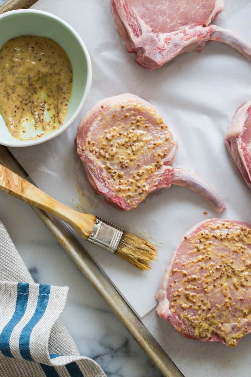 Raw pork chops that have been brushed with mustard with a bowl and a brush.