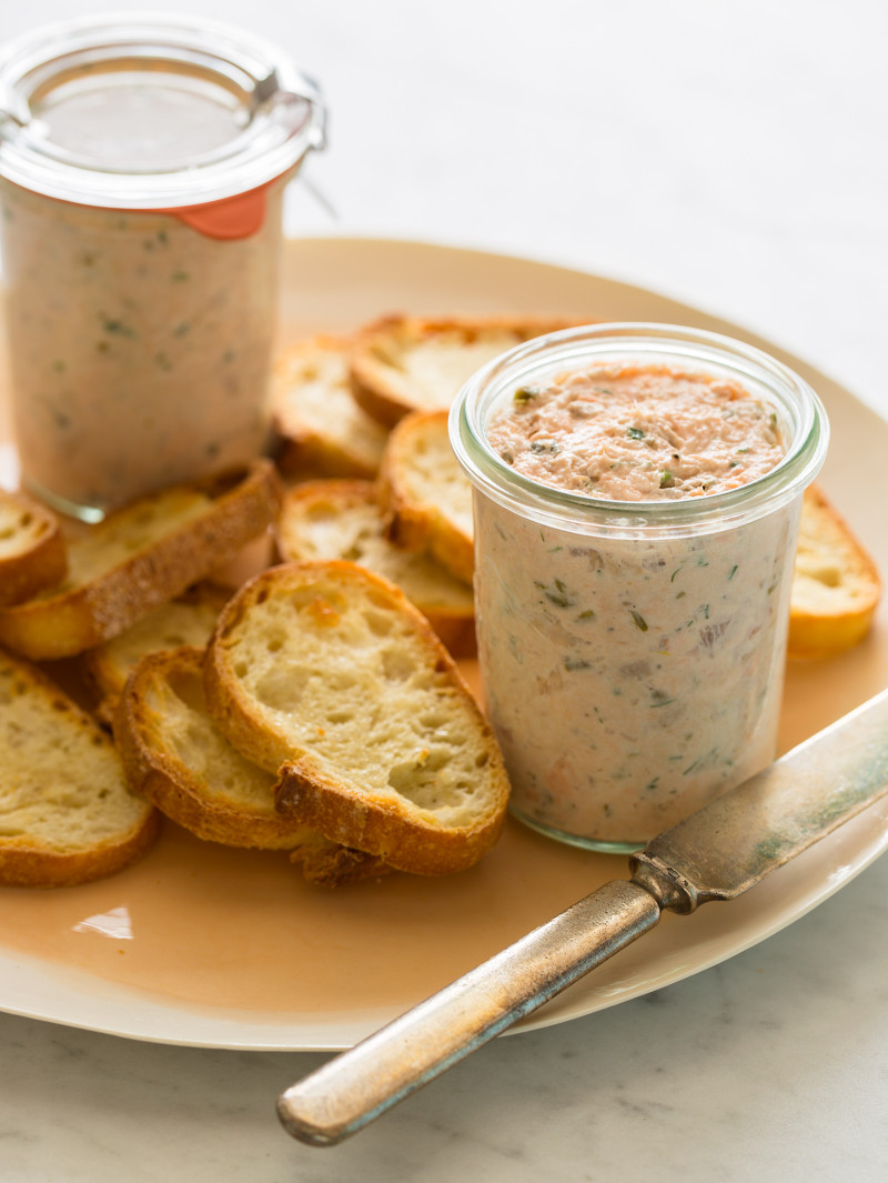 Glass jars of salmon rillettes on a plate with crostini and a knife.