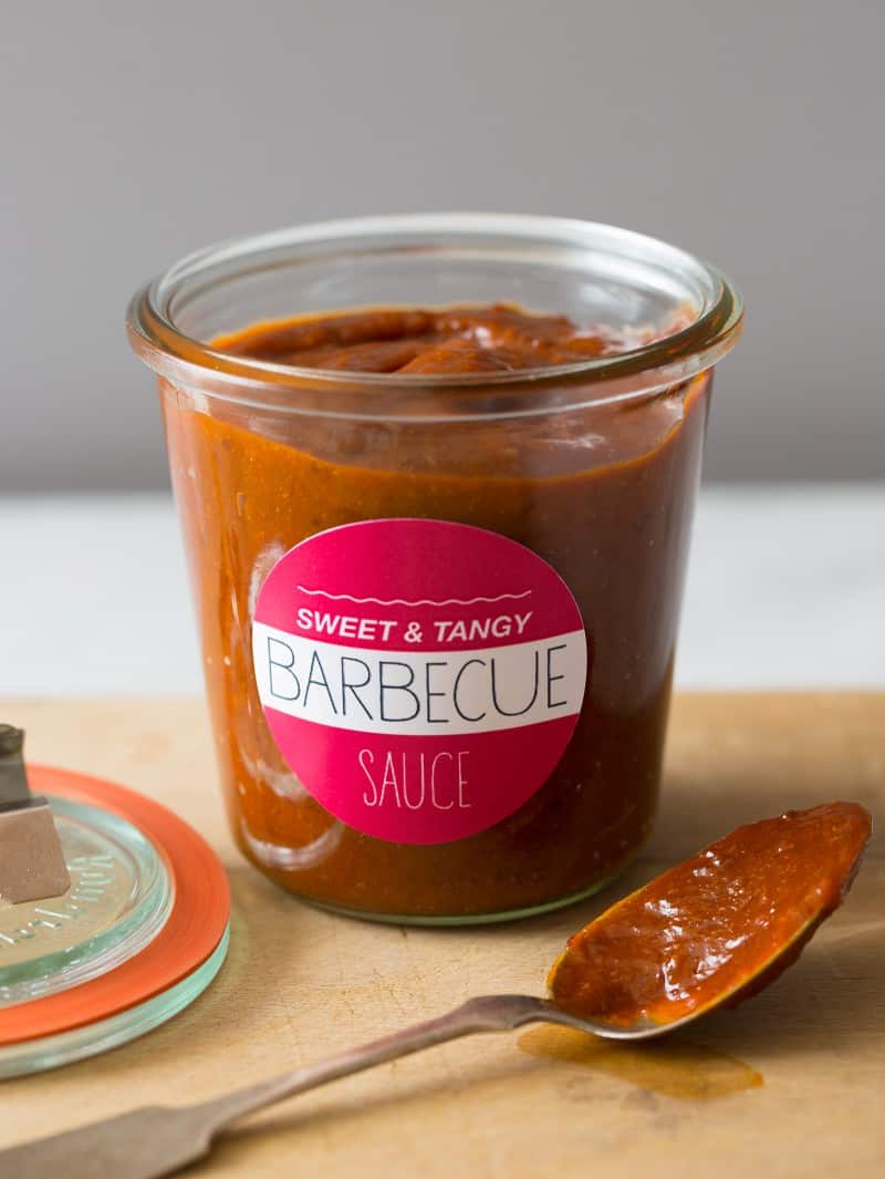 A labeled glass jar of sweet and tangy BBQ sauce, the lid, and a coated spoon.