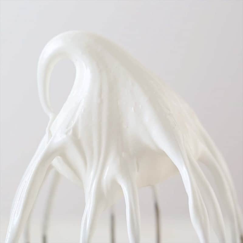 A close up of a medium stiff peak of meringue on the a mixer whisk.