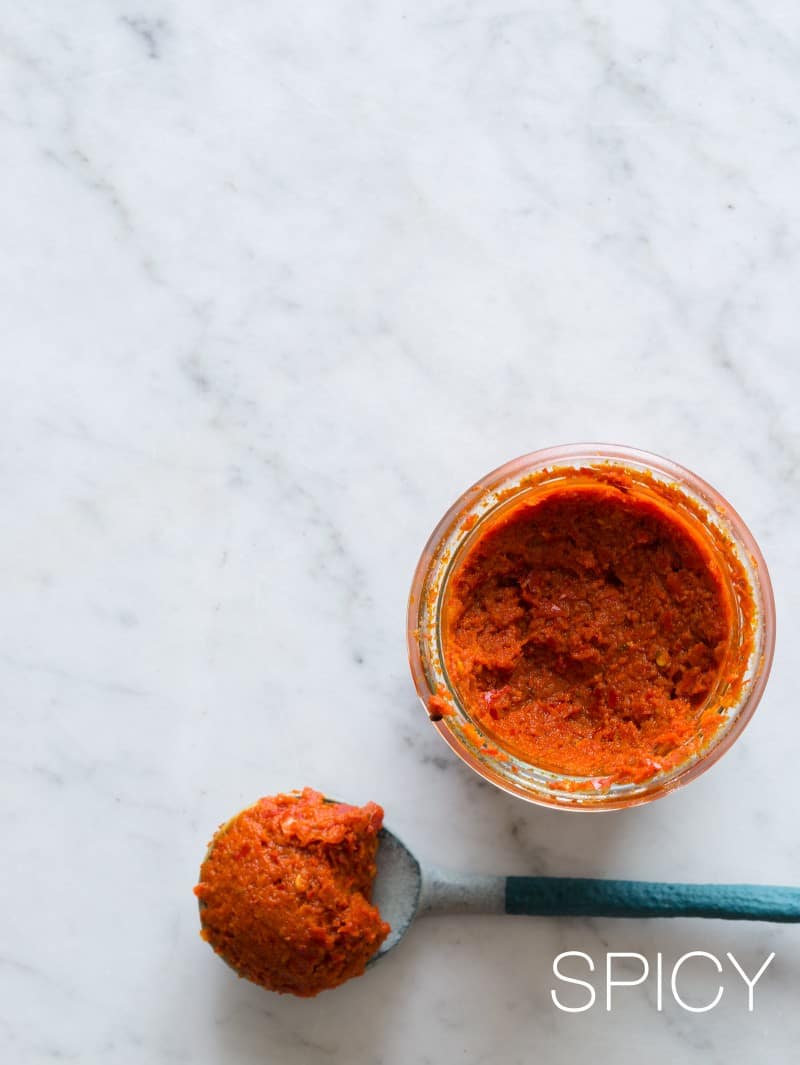 A close up of a blue spoonful of harissa next to a jar of spicy harissa.