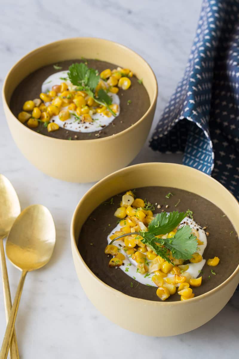 A close up of bowls of spicy black bean soup garnished next to spoons and linens.