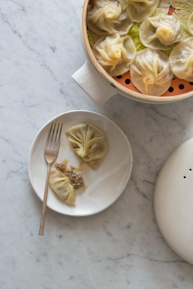 A steamer full of shanghai soup dumplings next to a plate of dumplings and a fork.