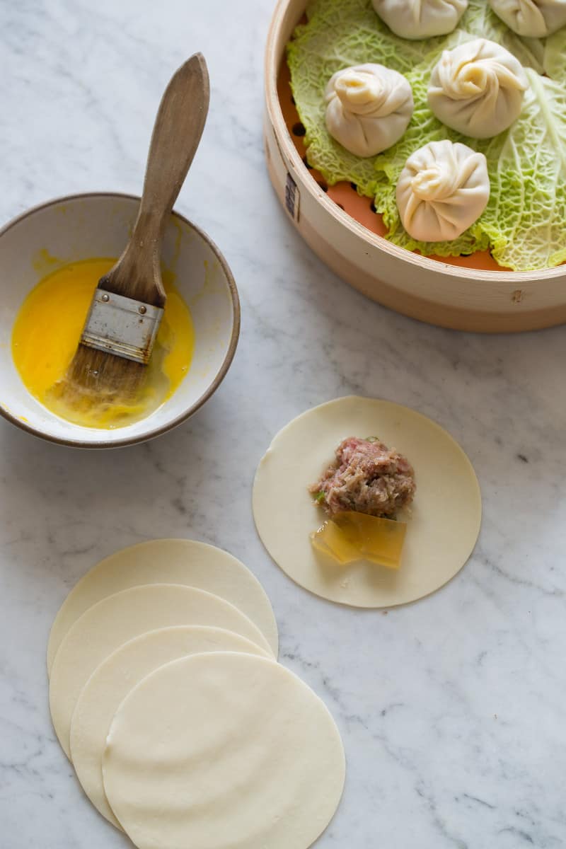 A bowl of egg with a brush and a dumpling being assembled next to a steamer.