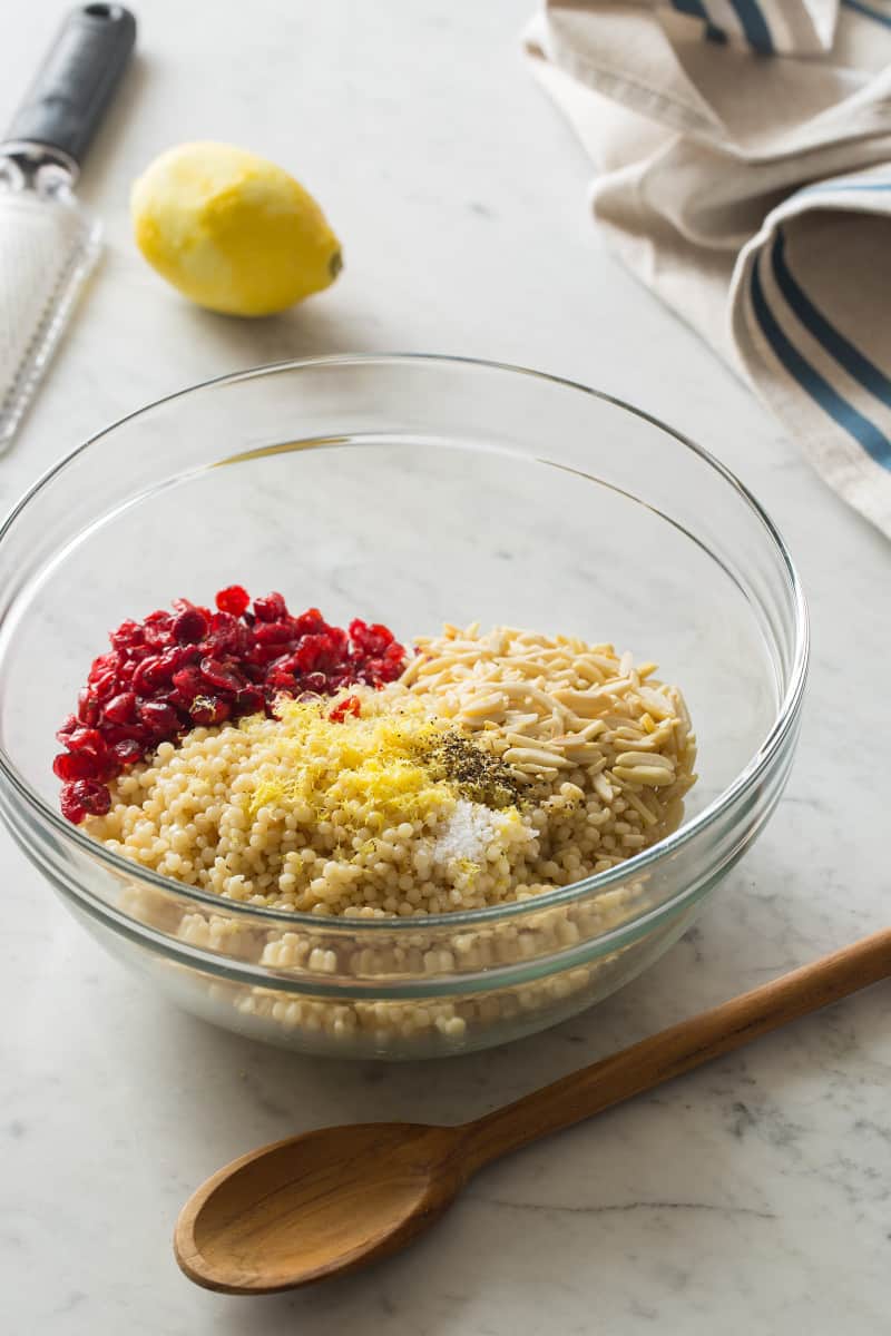 A clear mixing bowl of couscous ingredients with a wooden spoon.