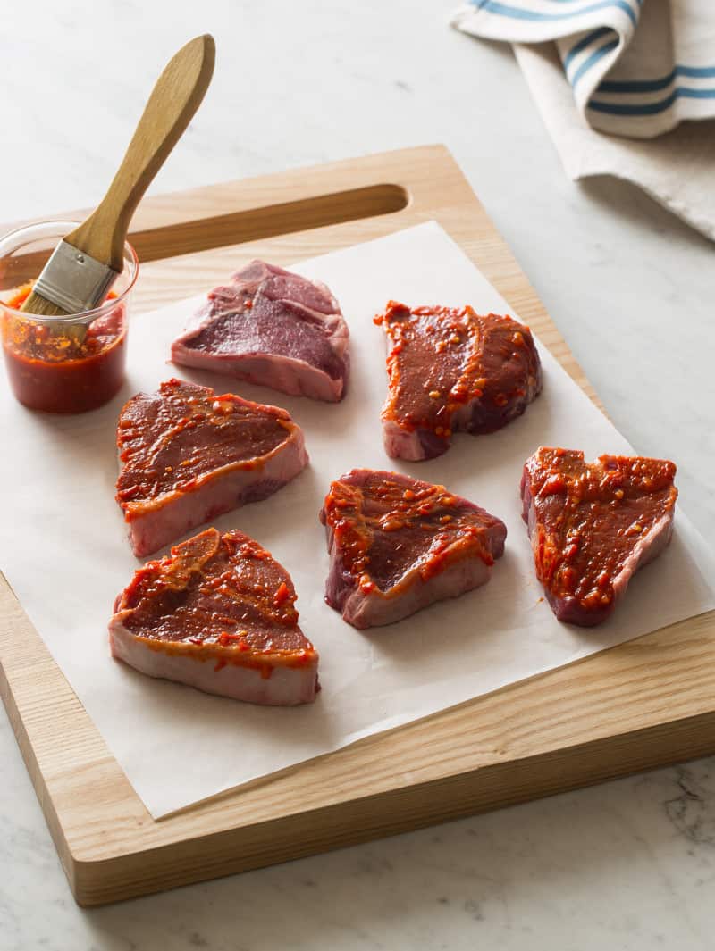 A wooden cutting board of raw lamb chops being brushed with sauce.