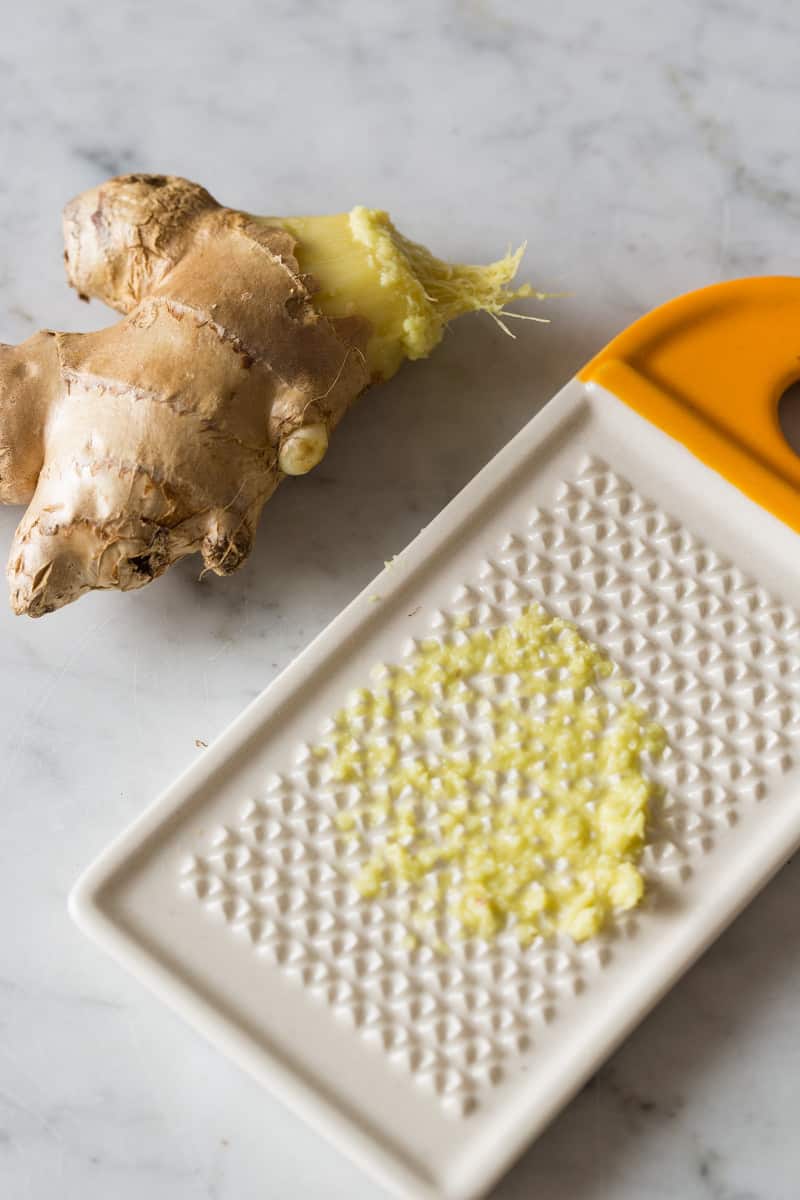 A close up of a ceramic grater, some fresh grated ginger, and a piece of ginger.