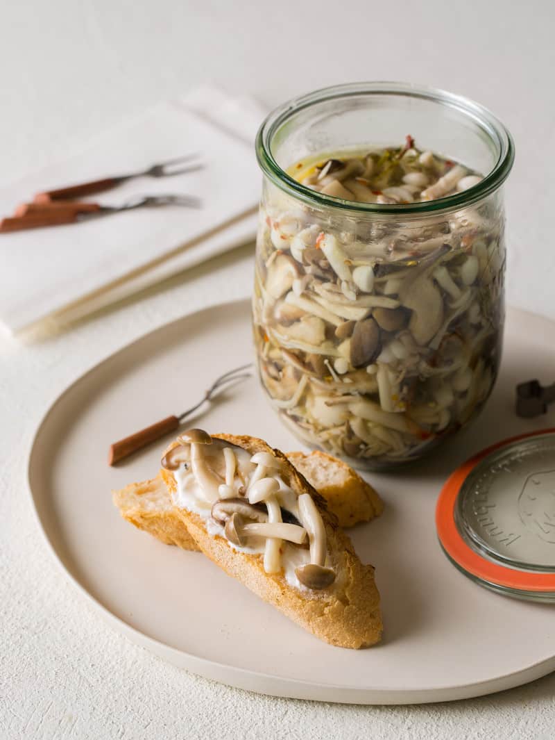 A plate with an open jar of pickled mushrooms with some mushrooms on bread.