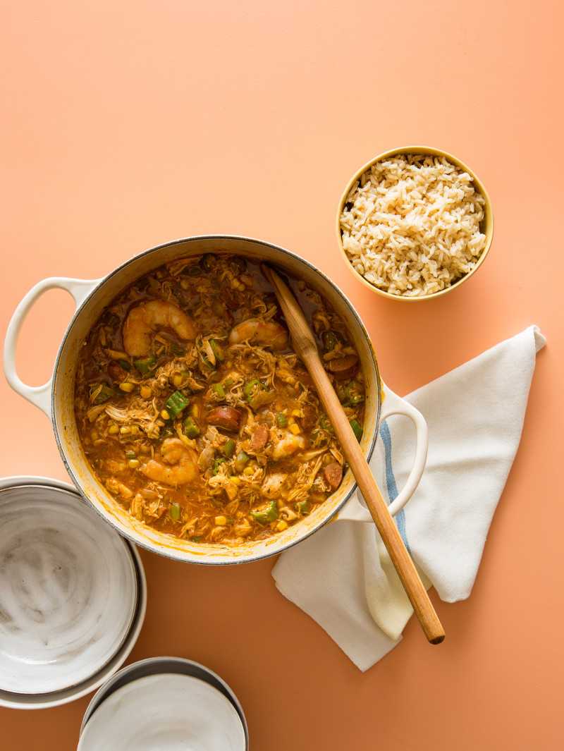 A pot of chicken and andouille gumbo with a bowl of rice and a wooden spoon.