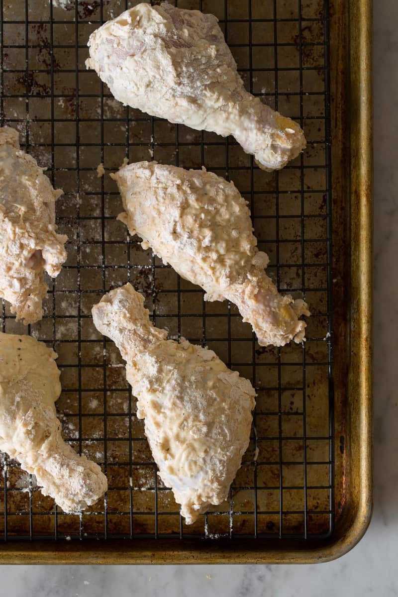 A close up of coated chicken drumsticks ready to be fried on a wire rack.