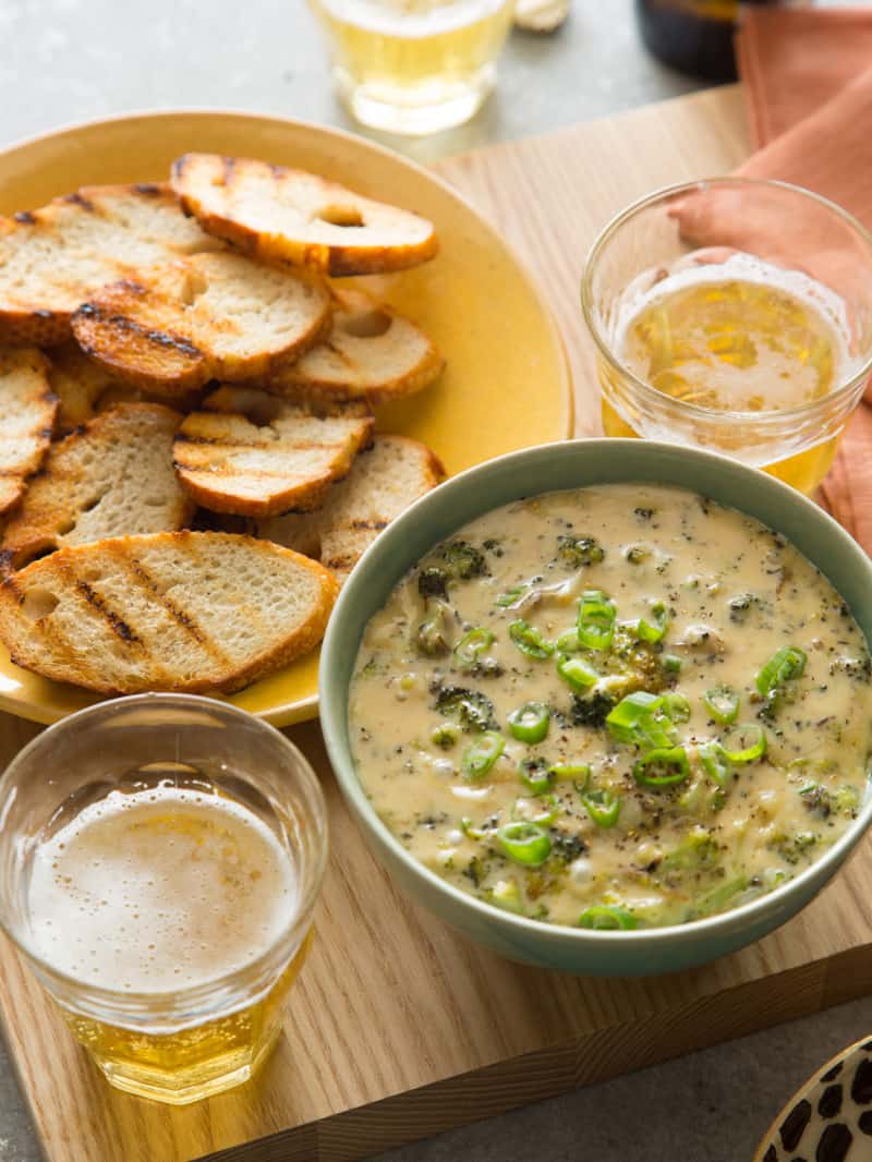 A bowl of roasted broccoli and white cheddar queso fundido with crostini and drinks.