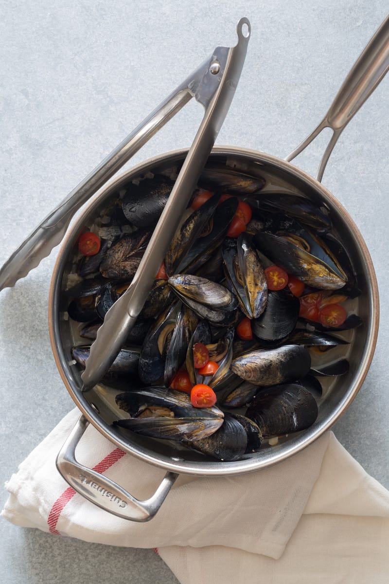 White wine steamed mussels in a pan with cherry tomatoes and tongs.