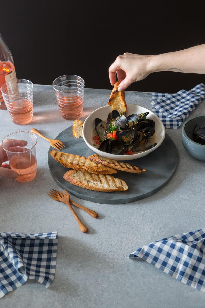 A person holding bread in a bowl of white wine steamed mussels with forks and drinks.