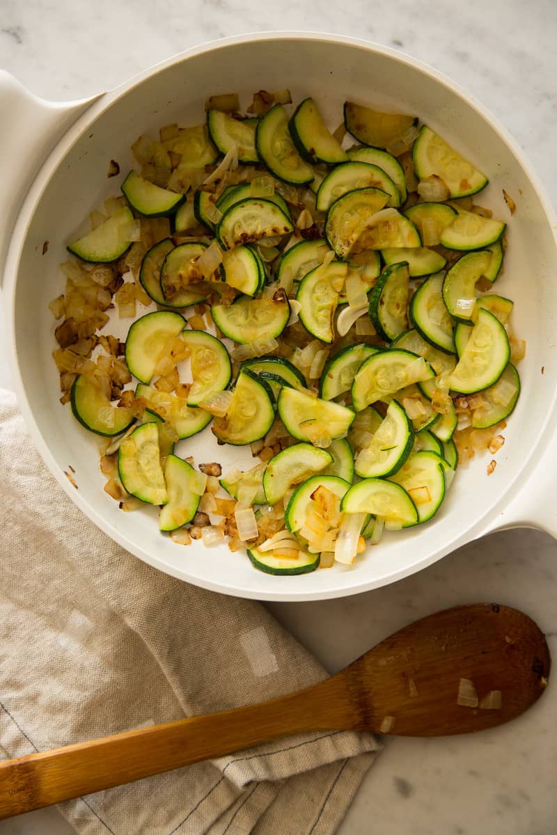 Sautéed onions and zucchini in a white pot with a wooden spoon.
