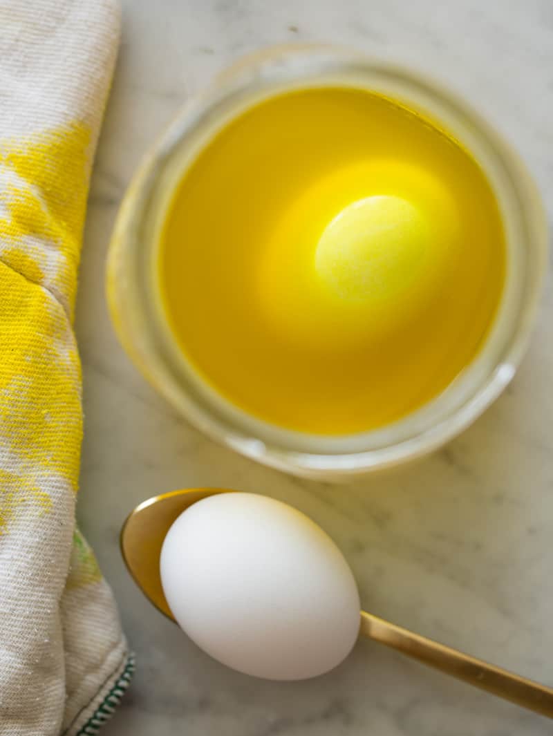 An egg in a bowl of yellow dye next to a bare egg on a spoon.