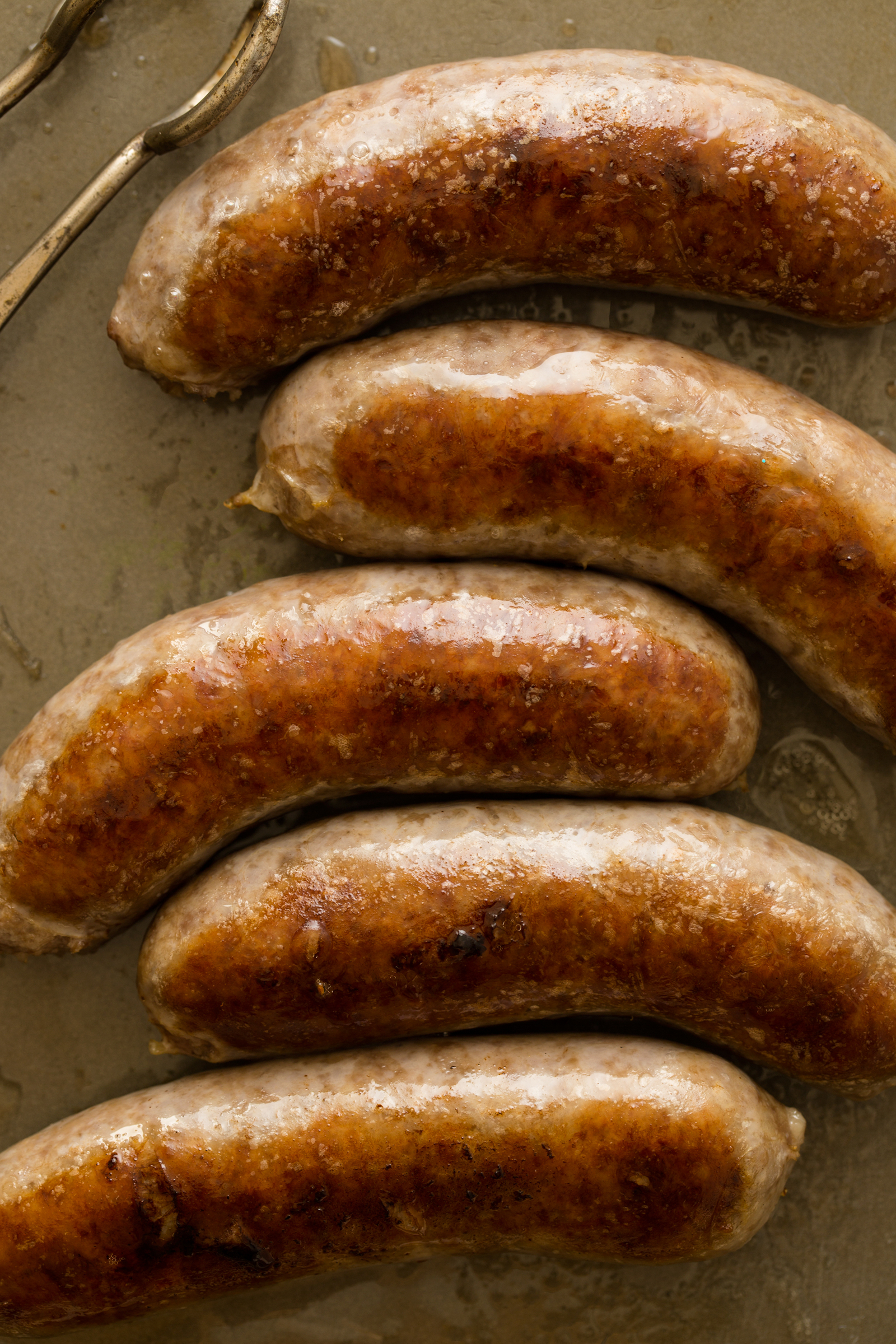 Freshly grilled bratwursts on a baking sheet next to tongs. 