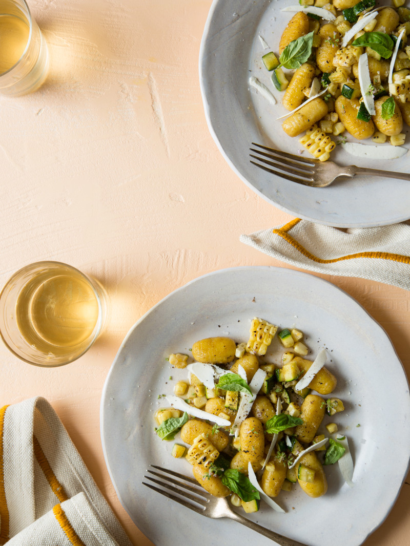 Summer gnocchi with charred sweet corn and zucchini on plates with forks and drinks.