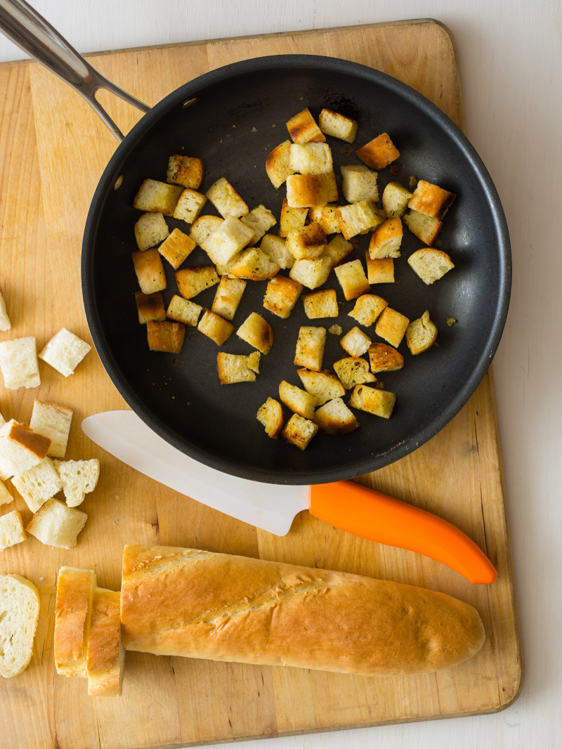 A loaf of bread cut into cubes with a knife and a pan of finished croutons.