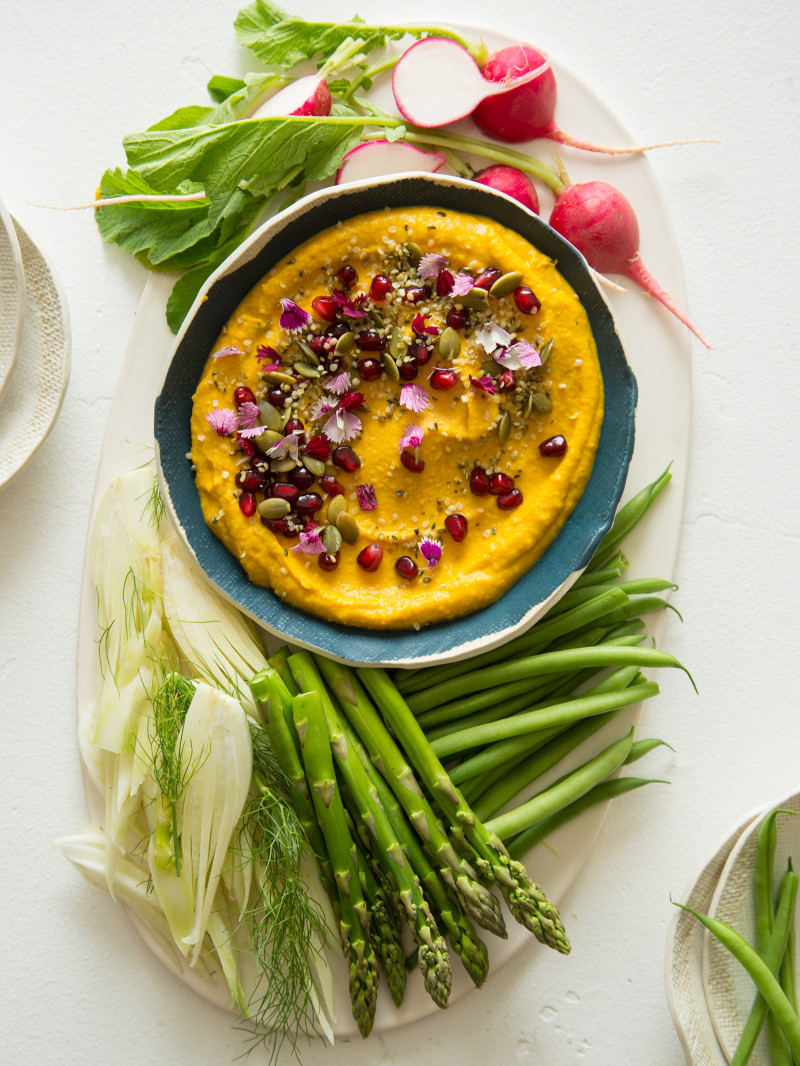 A bowl of roasted pumpkin hummus on a platter with a variety of veggies.