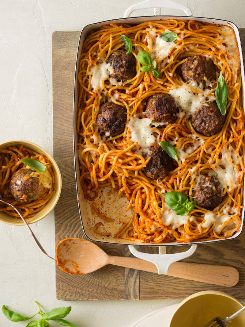 Baked bucatini and meatballs in a baking dish with a bowl served and a wooden spoon.