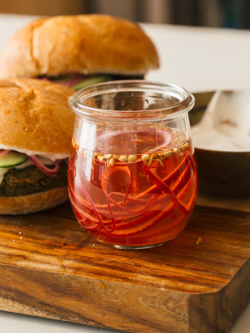 Falafel burgers on a wooden cutting board next to a jar of pickled red onions.