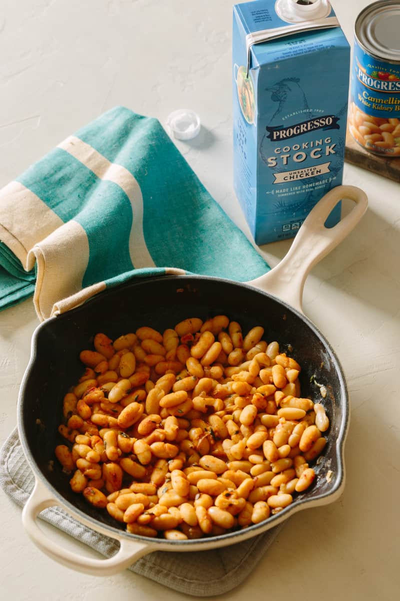 White beans in a skillet next to a box of chicken stock and a napkin.