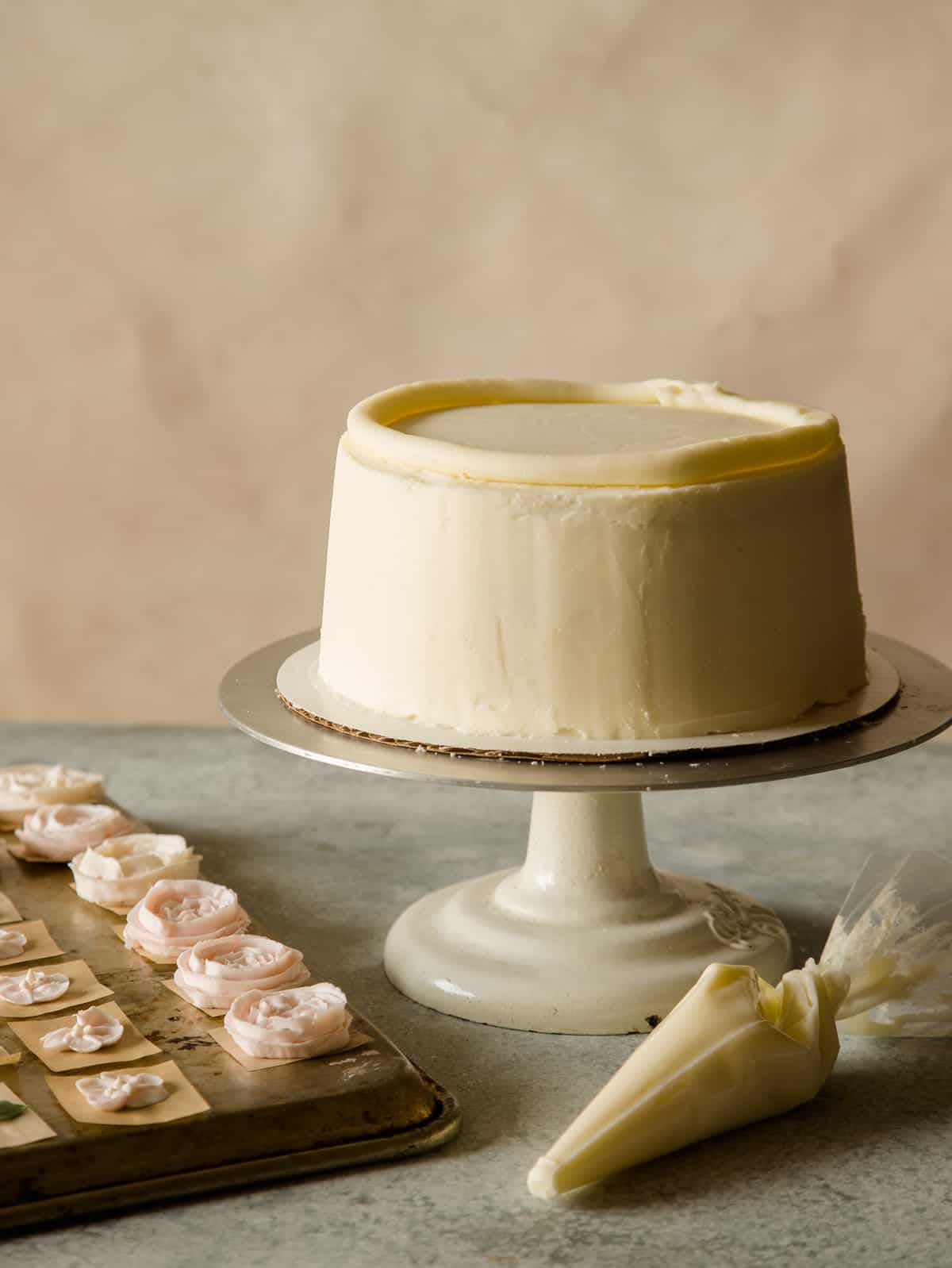 A frosted but undecorated cake on a cake stand with floral decor next to it.