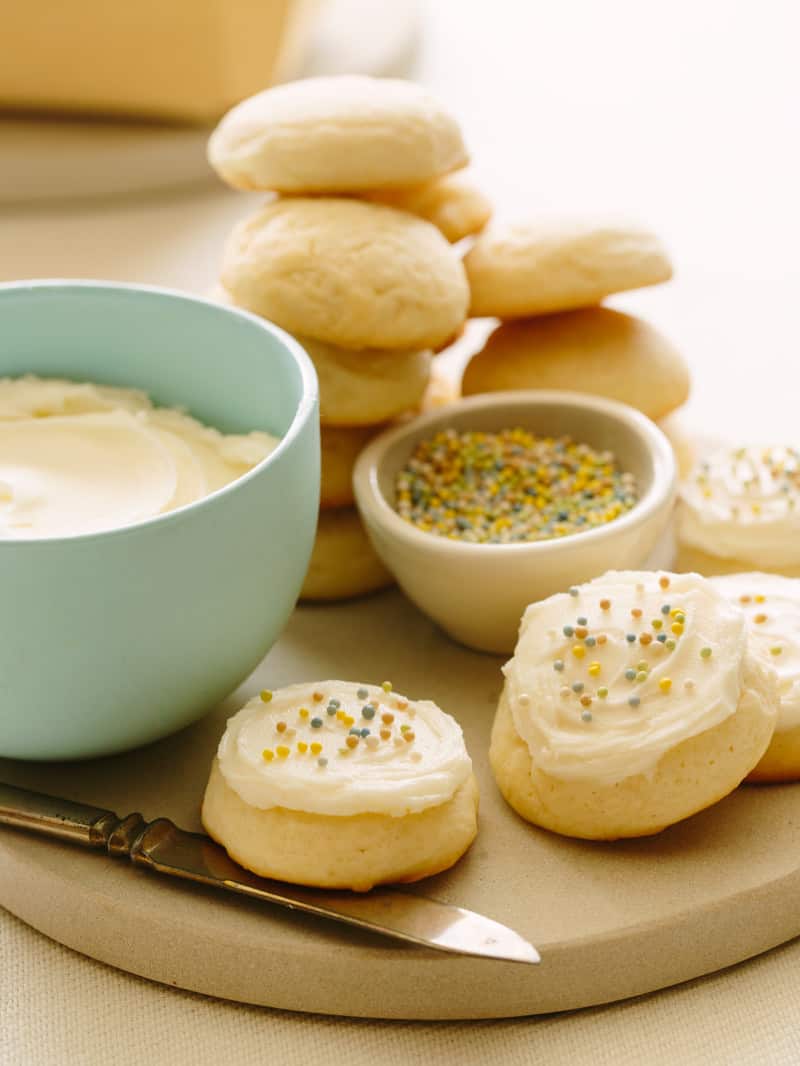 Soft and fluffy sugar cookies next to a bowl of vanilla frosting and sprinkles.