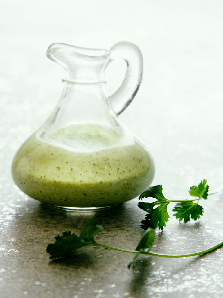 A close up of a glass pitcher of cilantro ranch dressing with a sprig of fresh cilantro.