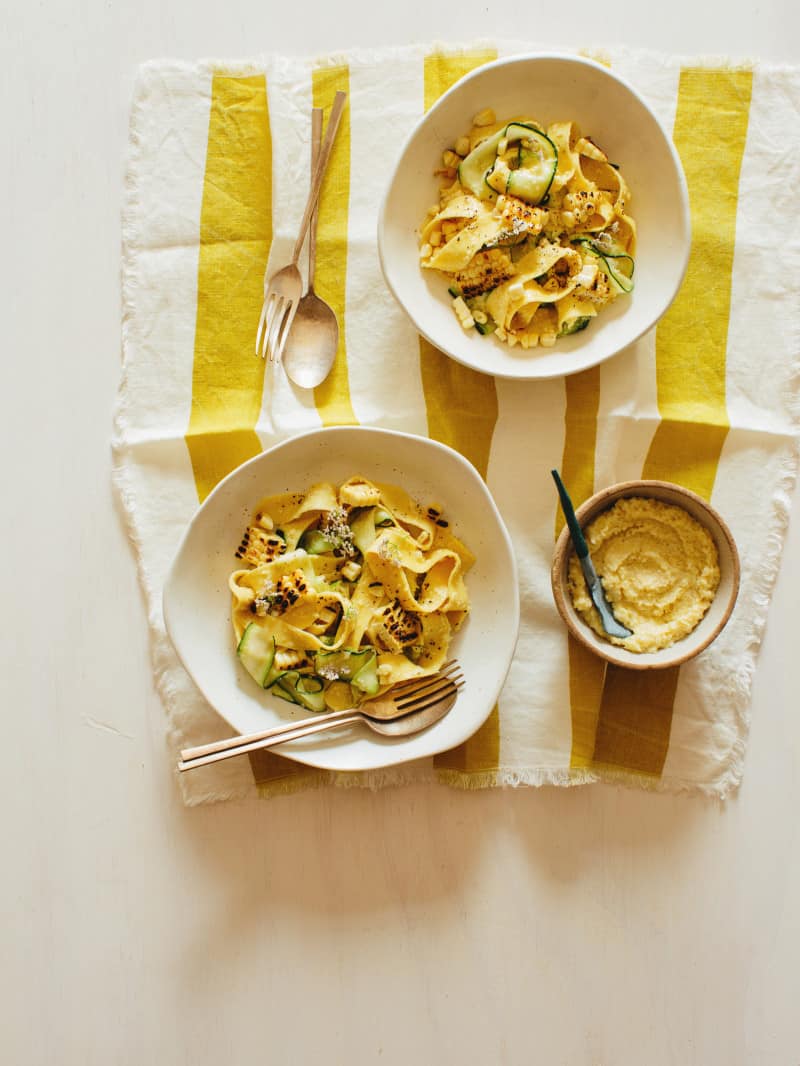 Bowls of sweet corn pesto with pappardelle and zucchini noodles on linens with forks and spoons.