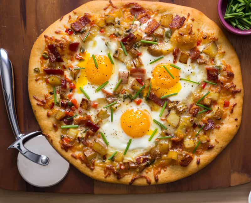 A breakfast pizza on a wooden cutting board with a pizza cutter wheel.