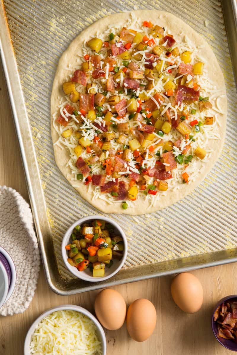 Uncooked breakfast pizza on a sheet pan with a bowl of potato mixture on the side.