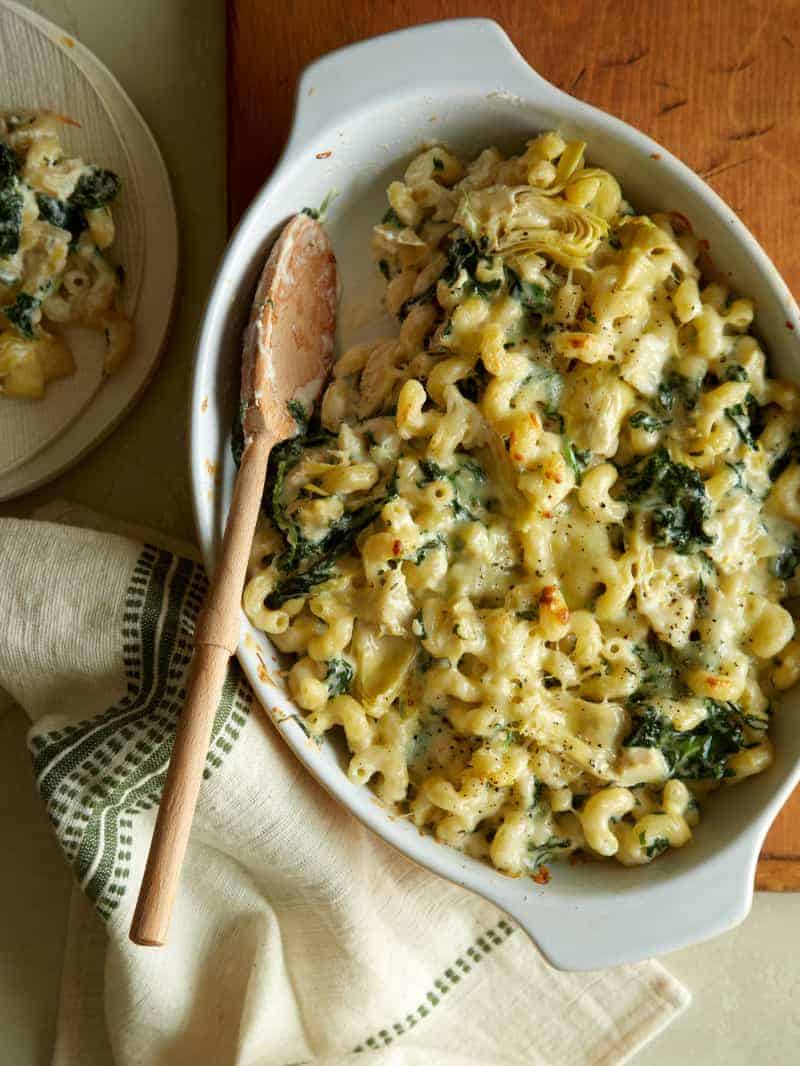 An oval baking dish with spinach artichoke mac and cheese with a wooden spoon.