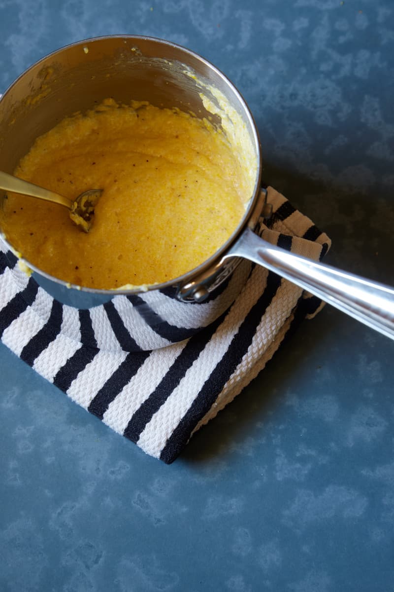 White cheddar polenta in a pot with a spoon sitting on a hand towel.
