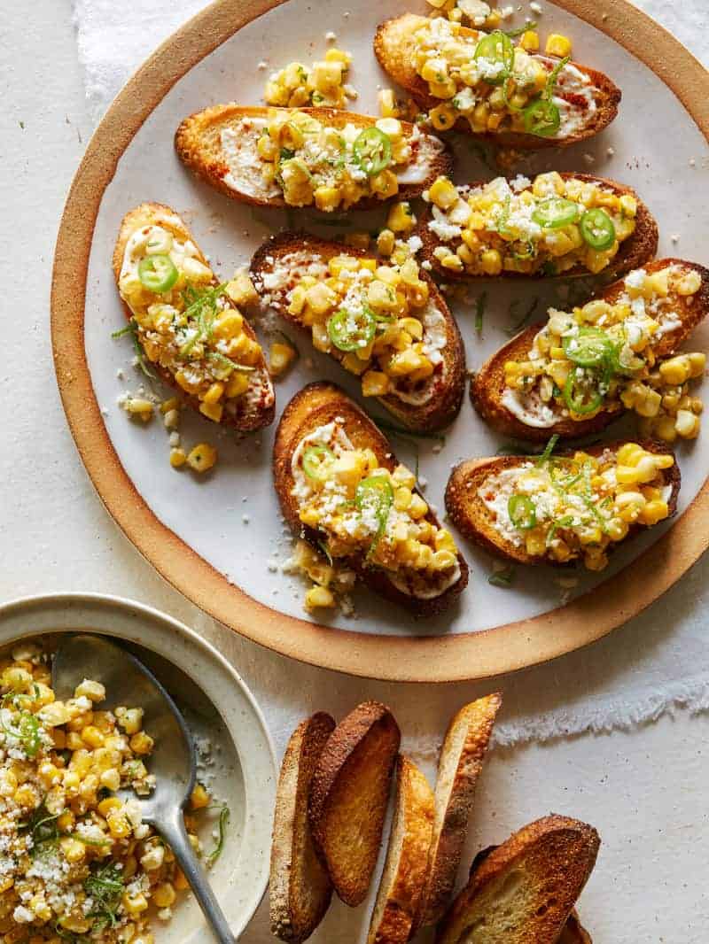 A plate of elote bruschetta with toppings and bread on the side.