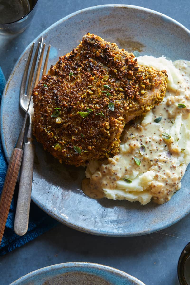 Pistachio crusted pork chop, partially sliced on mashed potatoes with a fork and knife.