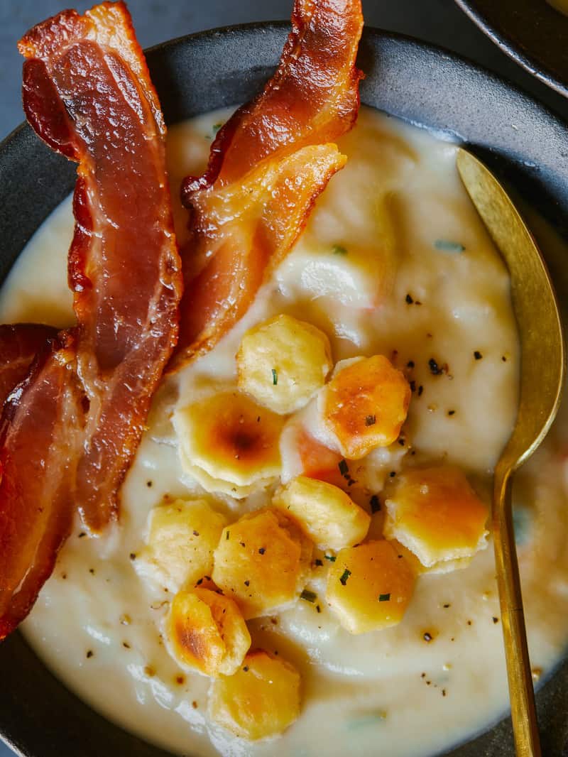 A close up of creamy vegan cauliflower chowder with crackers, bacon and a spoon.