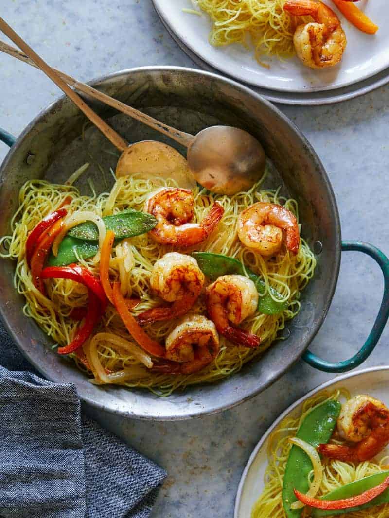 A pot of singapore noodles with shrimp and peppers and serving spoons.