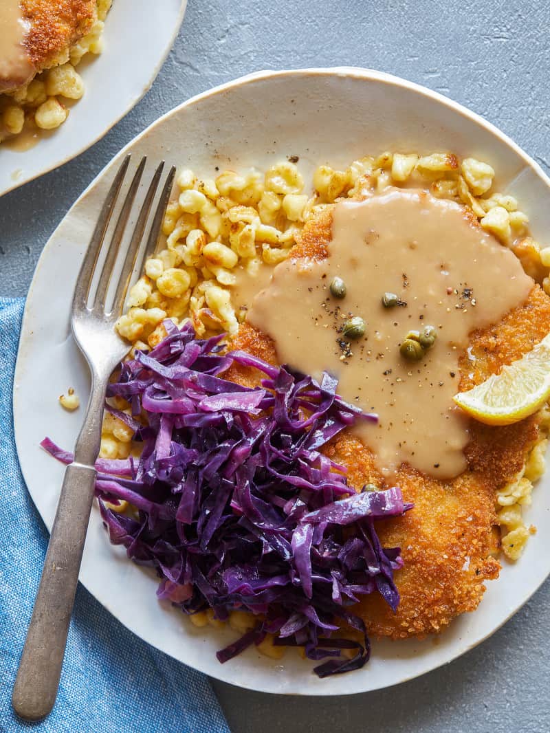 Schnitzel over buttered spaetzle with sweet and sour cabbage, lemon wedges and a fork.