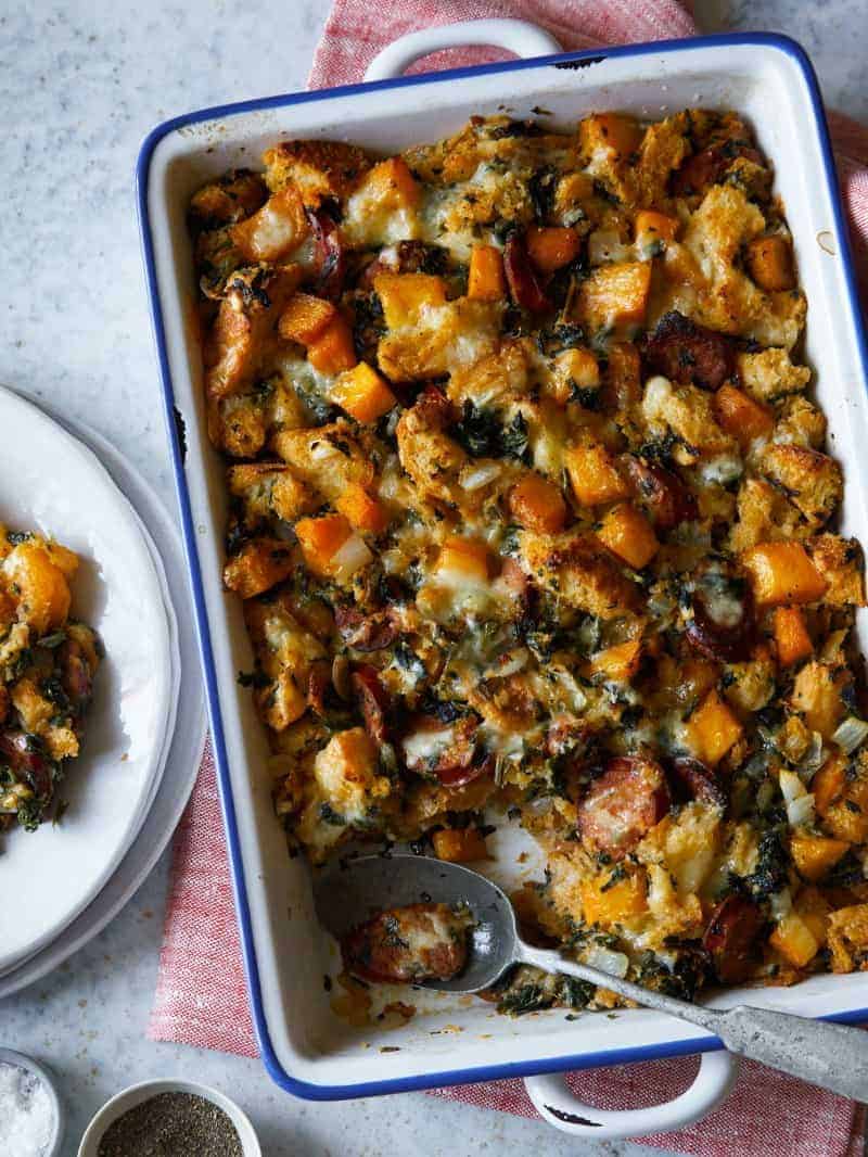 A baking dish of butternut squash sage stuffing with plates and a spoon.