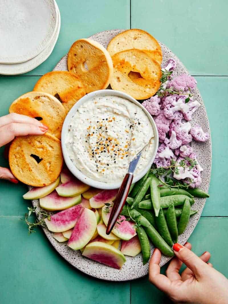 Everything seasoning and caramelized onion dip on a platter with bagels and veggies.