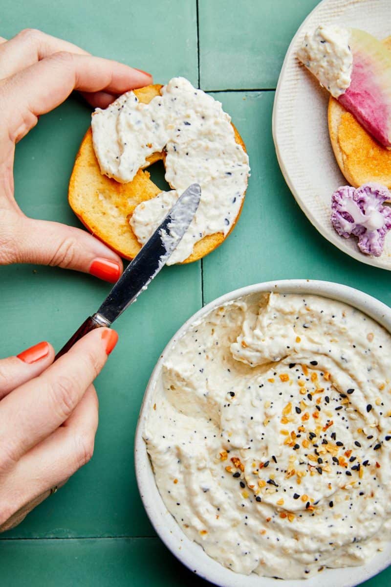 A woman spreading everything seasoning and caramelized onion dip on a bagel.