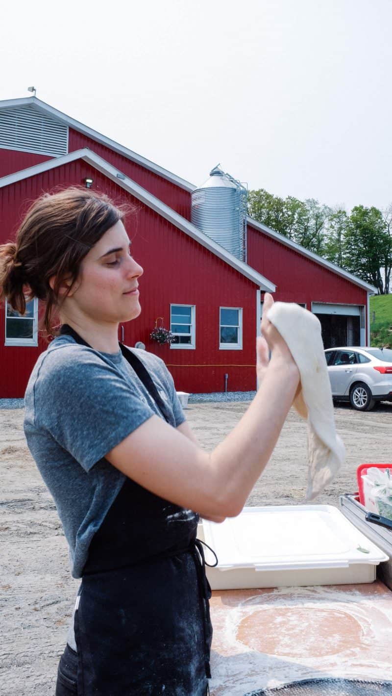 A woman holding pizza dough in front of a farm.