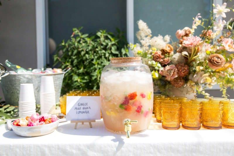 A glass drink dispenser with lemonade next to glasses and a sign.