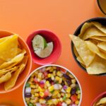 A bowl of grilled corn salsa next to bowls of tortilla chips and lime wedges.