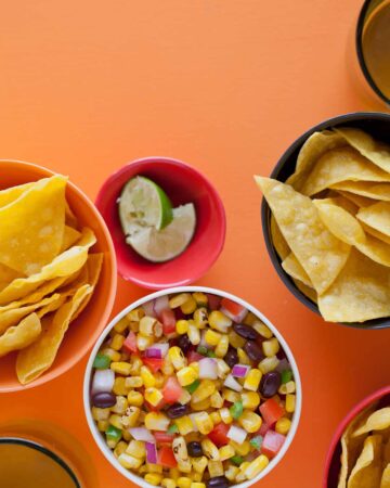 A bowl of grilled corn salsa next to bowls of tortilla chips and lime wedges.