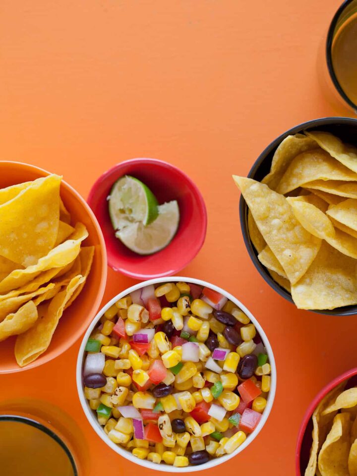A bowl of grilled corn salsa next to bowls of tortilla chips and lime wedges.